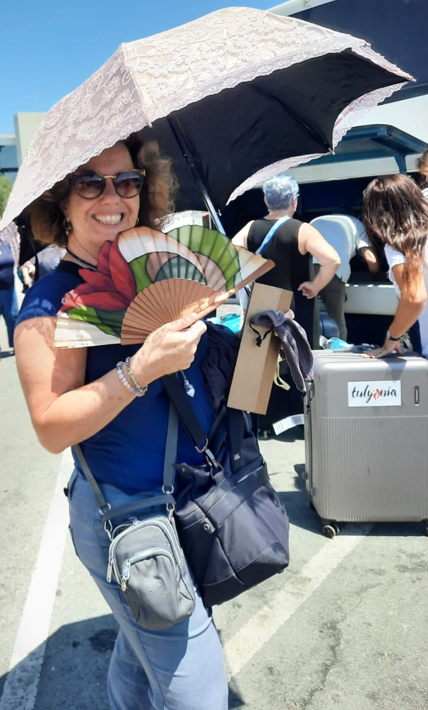 Gaby holding a fan with a floral design, also holding a bottle of body oil and an umbrella, with other people in the background. Tulynia Larnaka