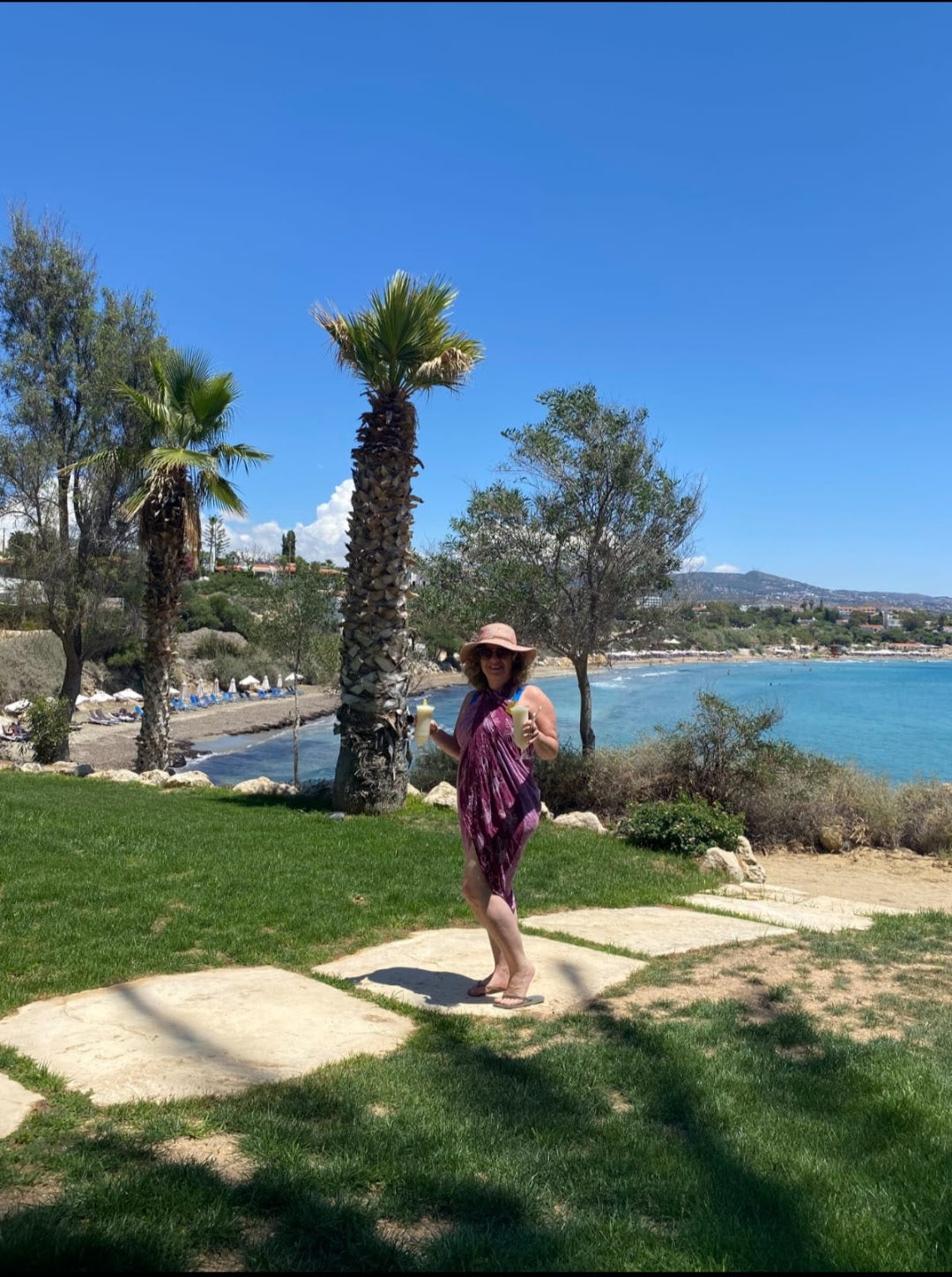 A person standing on a grassy area near a palm tree with a body of water and more palm trees in the background under a clear sky. Gaby holding Pina Colada in Paphos, Cypruss