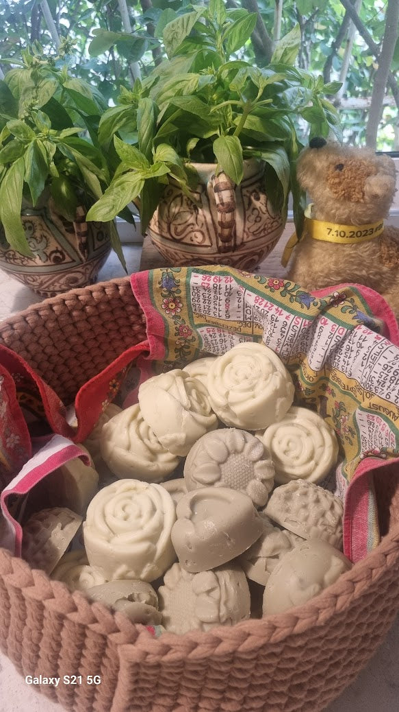 A basket filled with bars of organic soap with a distinctive blue lotus shape, placed in a setting with a plant and a candle, suggesting a spa-like atmosphere. Basil Decoration an October 7 yellow Ribbon.
